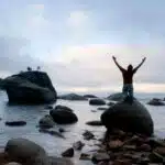 person on a shoreline kneeling on a rock with their arms stretched wide